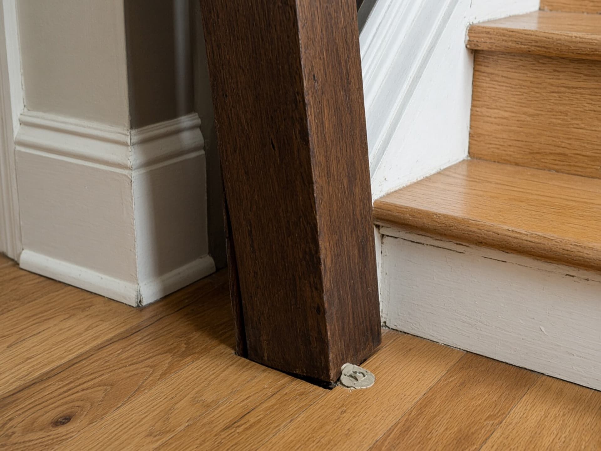 A newel post surface-fixed through a finished floor rather than anchored into structure, with the joint already opening at the base.